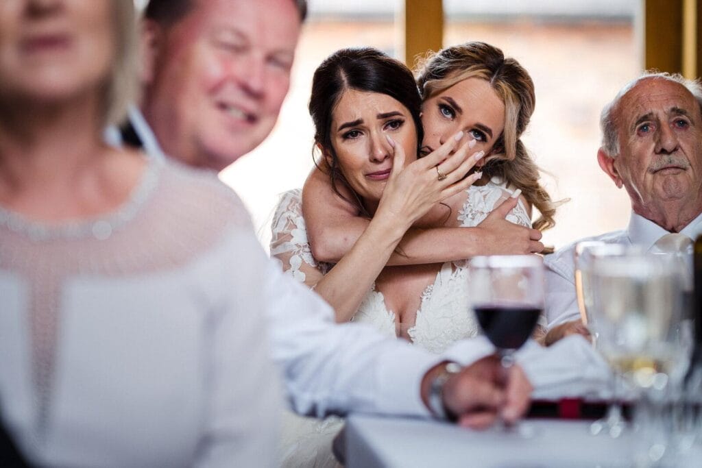 Emotional bride embracing a bridesmaid during a heartfelt wedding ceremony moment surrounded by family and guests.