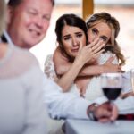 Emotional bride embracing a bridesmaid during a heartfelt wedding ceremony moment surrounded by family and guests.
