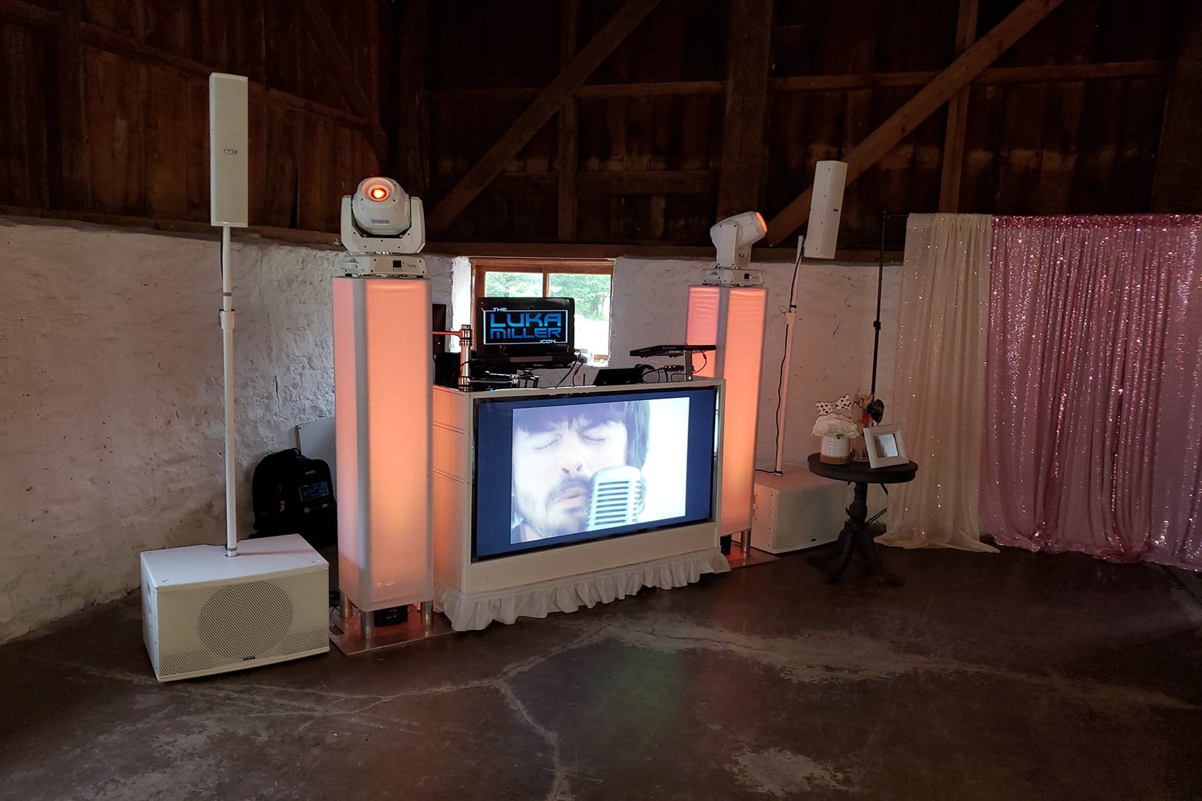 White wedding DJ and video booth setup with speakers and lighting in a rustic barn